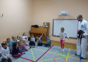 dzieci siedzą skrzyżnie i patrzą jak dziewczynka skacze z koła do koła obok instruktora