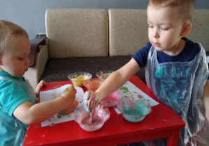 bracia siedzą przy czerwonym, małym stoliku, na którym stoja pojemniki z kolorowymi farbami i malują rękami