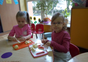 dwie dziewczynki siedzą przy stoliku i kroją na desce owoce