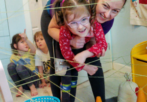 mama trzyma córkę na rękach przed labiryntem z żółtych nitek a a z tyłu siedzą dwie córki