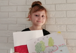 dziewczynka stoi na tle białej ściany z cegiełek i trzyma flagę polską oraz ilustrację parku miejskiego ze stawem, ławkami, wierzbami i kaczkami