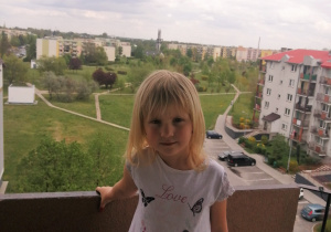dziewczynka stoi na balkonie swojego domu a za nią roztacza się widok na osiedle bloków, parking i łąkę