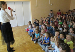 pani mówi do mikrofonu a dzieci siedzą na dywanie i sluchają