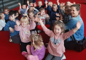 dzieci z panią siedzą w grupce na holu w teatrze i machają