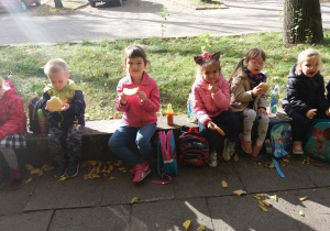 dzieci siedzą na dworze, na murku i jedzą bułki z serem