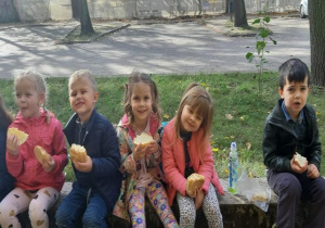 dzieci siedzą na dworze, na murku i jedzą bułki z serem