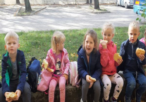 dzieci siedzą na dworze, na murku i jedzą bułki z serem