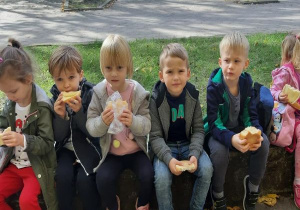dzieci siedzą na dworze, na murku i jedzą bułki z serem