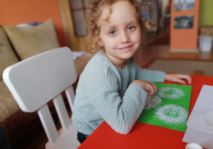 w domu, dziewczynka siedzi przy stole i stempluje białe dmuchawce na zielonej kartce