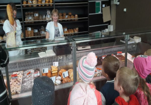 pani pokazuje podłużny chleb z makiem dzieciom a obok stoi druga sprzedawczyni