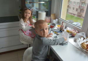 rodzeństwo siedzi w domu, w kuchni, na stołkach i przygotowuje deser dla mamy z różnych składników