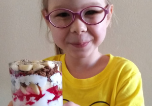 dziewczynka trzyma w rękach szklankę z deserem dla mamy złożonym z bitej śmietany, czerwonej galaretki, orzechów i czekoladowych chrupek