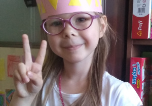 dziewczynka stoi i ma na głowie różowy pióropusz z naklejonymi żółtymi trójkątami (opaska) oraz cienkimi, czerwonymi paskami paieru (pióra)wykonany przez siebie z kolorowego papieru