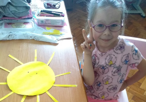 dziewczynka siedzi przy stole, na którym leży słoneczko wycięte z żółtego papieru w raz z promieniami w kształcie cienkich pasków