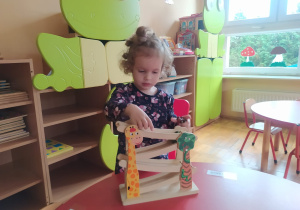 dziewczynka stoi przy czerwonym stoliku i bawi się zjeżdżalnią dla małych aut a w tle stoją szafki z wielka zieloną żabą