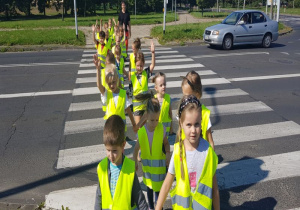 dzieci ubrane w kamizelki odblaskowe w kolorze cytrynowym przechodzą przez jezdnię po pasach