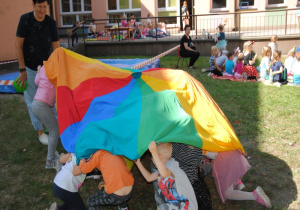 w ogrodzie podczas zabawy chustą na znak stojącej obok nauczycielki dzieci chowają sie pod chustę