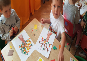 dzieci przy stoliku robią wydzierankę z kolorowego papieru (liście) i naklejają na kontur drzewa