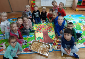 dzieci z gr. III siedzą w sali wokół wspólnie wykonanej pracy plastycznej - drzewo z występlowanymi farbą, połówkami jabłek a obok leżą dwie tace: jedna z naturalnymi jabłkami a druga z suszonymi jabłkami