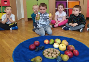 dzieci siedzą w kole, w środku leży granatowa chusta z owocami a chłopiec trzyma gruszki
