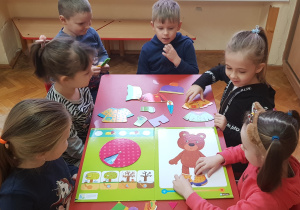 dzieci siedzą przy stoliku i ubierają misia