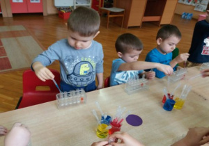 dzieci siedzą przy stolikach i pipetami nabierają kolory i w lewaja do probówek