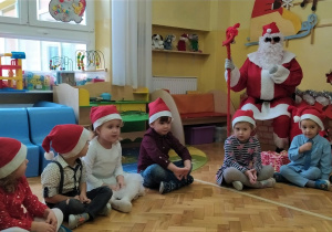 Mikołaj siedzi obok choinki z laską w dłoni a przed nim siedzą dzieci z paczkami