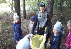 nauczycielka trzyma żółty worek a dzieci wrzucają do niego znalezione w lesie śmieci