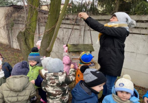 dzieci stoją a pani zawiesza słoninkę przy karmniku