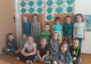 dzieci z grupy V ubrane w kolory ziemi stoją przed tablicą ze swoimi pracami o Ziemi