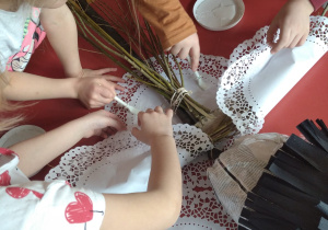 dzieci przy stoliku przyklejają na rękach Marzanny z gałęzi papierowe, białe, duże, ażurowe serwetki jako jej bluzkę