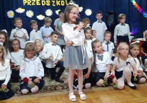dzieci z grupy III na uroczystości pożegnania przedszkola - dziewczynka recytuje wiersz na tle innych dzieci ze swojej grupy