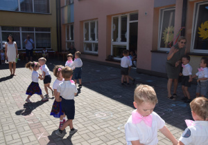 dzieci z grupy I tańczą w parach na tarasie przedszkolnym trzymając się za ręce, ubrane w białe koszulki z różowymi muchami