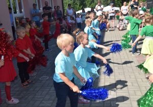 dzieci z grupy IV - tańczą z pomponami, ubrani w trzech kolorach: zielonym, czerwonym i niebieskim ustawieni w rzędach