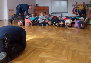 policjant demonstruje właściwe zachowanie dzieciom podczas ataku psa na człowieka a dzieci naśladują jego zachowanie, drugi policjant sprawdza ułożenie ciała dzieci