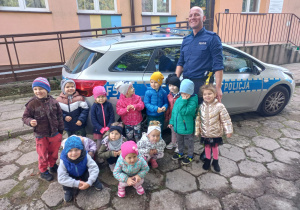 dzieci z grupy V stoją z policjantem obok radiowozu