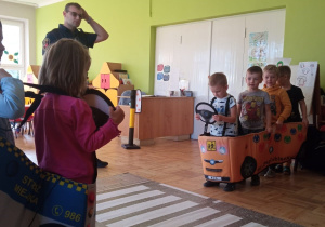 dzieci bawią się ze strażnikiem miejskim - dzieci ustawione w pojazdach po dwóch stronach przejścia dla pieszych - pasów ułożonych na podłodze sali
