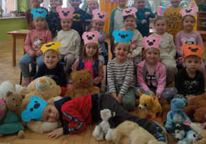 dzieci z grupy I pozują do zdjęcia grupowego w kolorowych opaskach misiów a u ich stóp leżą pluszaki a na nich jeden z kolegów
