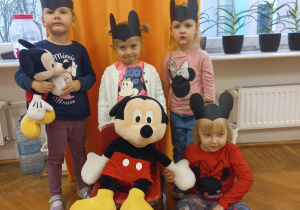 cztery dziewczynki stoją wokół dużej maskotki Myszki Miki siedzącej na krzesełku