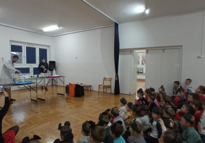 dzieci siedzą i przyglądają się występowi chemika