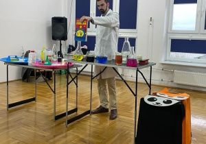 chemik pokazuje książkę dzieciom trzymając ją nad menzurkami wypełnionymi kolorowymi cieczami