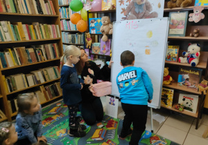 w bibliotece - dzieci siedzą i obserwują jak kolega rysuje misia na dużym, białym arkuszu papieru