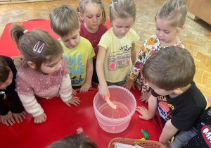 dzieci stoją wokół pojemnika z wodą a dziewczynka trzyma zapaloną świeczkę i leje wosk na wodę