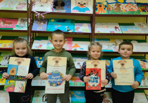 w bibliotece - dzieci z książkami i dyplomami na tle regału z pracami plastycznymi zgłoszonymi do konkursu plastycznego pod hasłem "Ja i mój pies"
