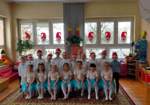 przedszkolaki z grupy III pozują do zdjęcia grupowego - dziewczynki w białych bluzkach i niebieskich, tiulowych spódniczkach a chłopcy w ciemnych spodniach, białych koszulach z niebieskimi muchami i czerwonych czapkach