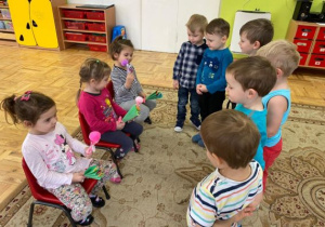 dziewczynki siedzą na krzesełkach i glądają swoje wachlarzyki a chłopcy stoją przed nimi
