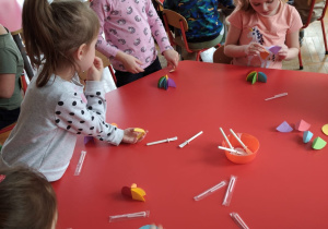 przedszkolaki przy stoliku wykonują parasolki z kolorowego papieru i słomki