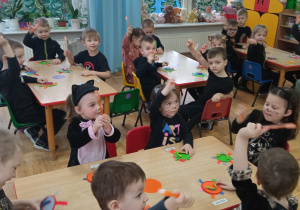 dzieci siedzą przy stolikach i pokazują swoje pomarańczowe szczoteczki unosząc je w górę