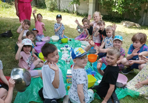 przedszkolaki machają siedząc w kole a obok stoi nauczycielka