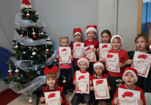 przy choince są dzieci w czerwonych strojach z grupy IV z wykonanymi podobiznami Mikołaja